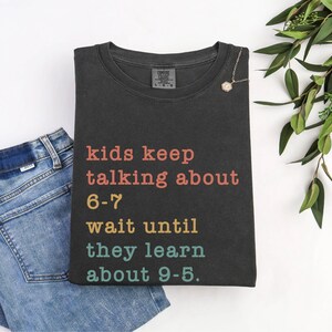 May include: Dark grey t-shirt with the text "kids keep talking about 6-7 wait until they learn about 9-5" in various colours. The shirt is folded next to a pair of blue jeans and a necklace.