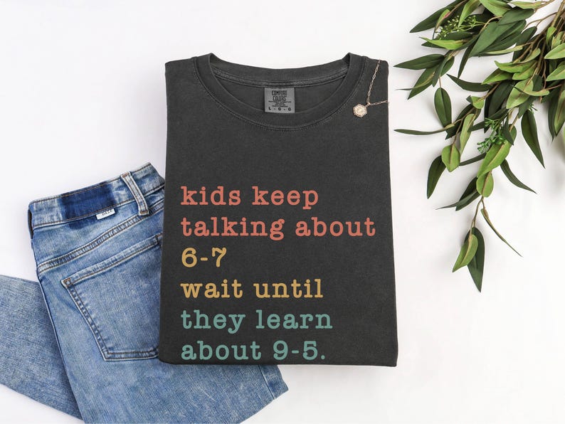 May include: Dark grey t-shirt with the text "kids keep talking about 6-7 wait until they learn about 9-5" in various colours. The shirt is folded next to a pair of blue jeans and a necklace.