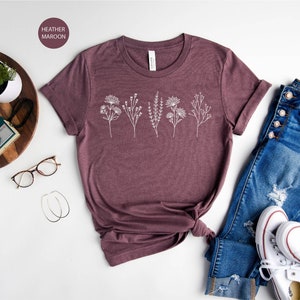 May include: A heather maroon t-shirt with a white floral design. The shirt is laid out on a white surface with a pair of jeans, a pair of white sneakers, and a wooden tray with a camera and a watch.