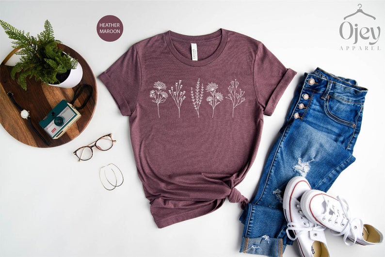 May include: A heather maroon t-shirt with a white floral design. The shirt is laid out on a white surface with a pair of jeans, a pair of white sneakers, and a wooden tray with a camera and a watch.