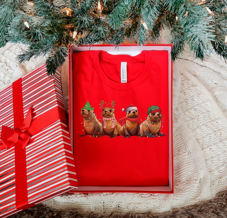 May include: Red t-shirt with a graphic of four seals wearing Christmas hats and lights. The seals are wearing a Santa hat, a reindeer hat, a green hat, and a green and red hat.