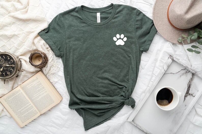 May include: A dark green t-shirt with a white paw print on the left chest. The t-shirt is knotted at the bottom and is lying on a white surface.
