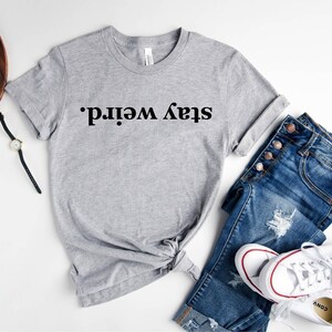 May include: A gray t-shirt with the text "Stay weird." printed in black. The shirt is styled with blue jeans and white sneakers.