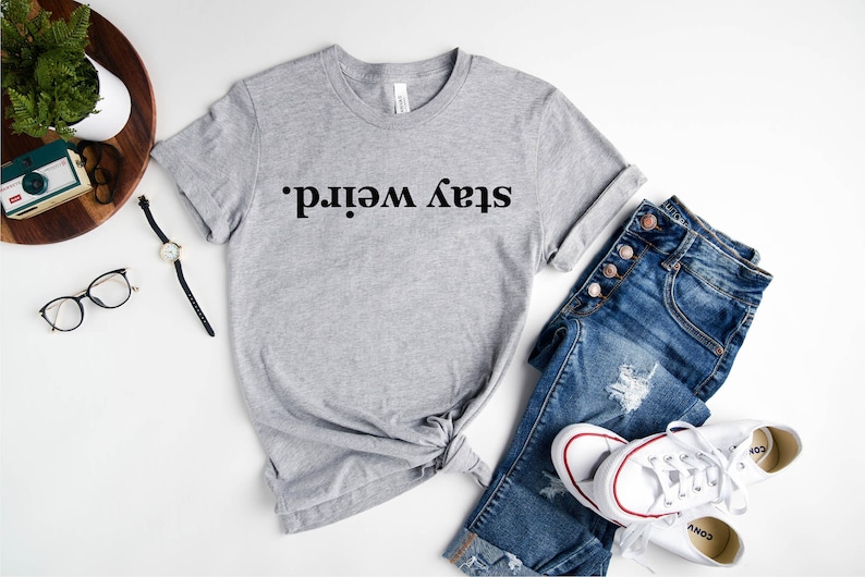 May include: A gray t-shirt with the text "Stay weird." printed in black. The shirt is styled with blue jeans and white sneakers.