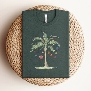 K&ouml;nnte beinhalten: Ein dunkelgr&uuml;nes T-Shirt mit einer Palme, die mit bunten Ornamenten geschm&uuml;ckt ist. Die Palme ist ein Weihnachtsbaum-Design.