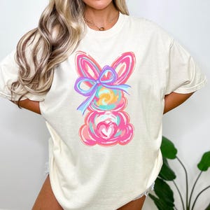 May include: A light beige t-shirt with a colorful abstract bunny design. The bunny has a pink body, blue ears, and a purple bow. The bunny is holding a pink heart.