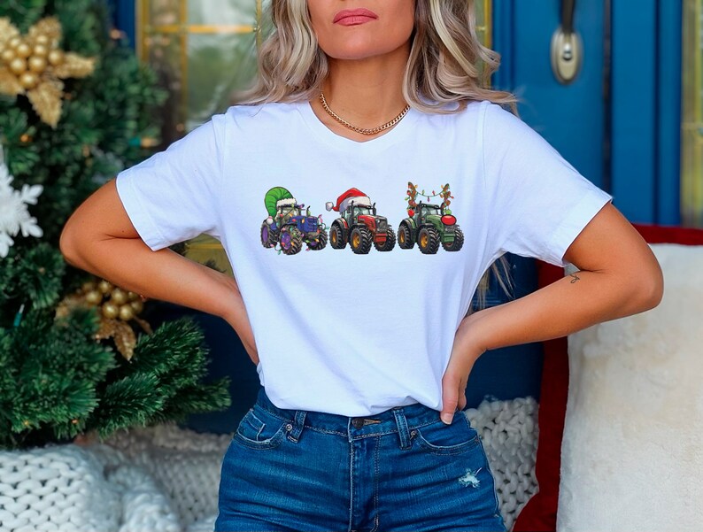 Peut inclure: T-shirt blanc avec un motif de trois tracteurs verts d&eacute;cor&eacute;s pour No&euml;l. Les tracteurs portent un chapeau de P&egrave;re No&euml;l, une couronne et un sapin de No&euml;l.