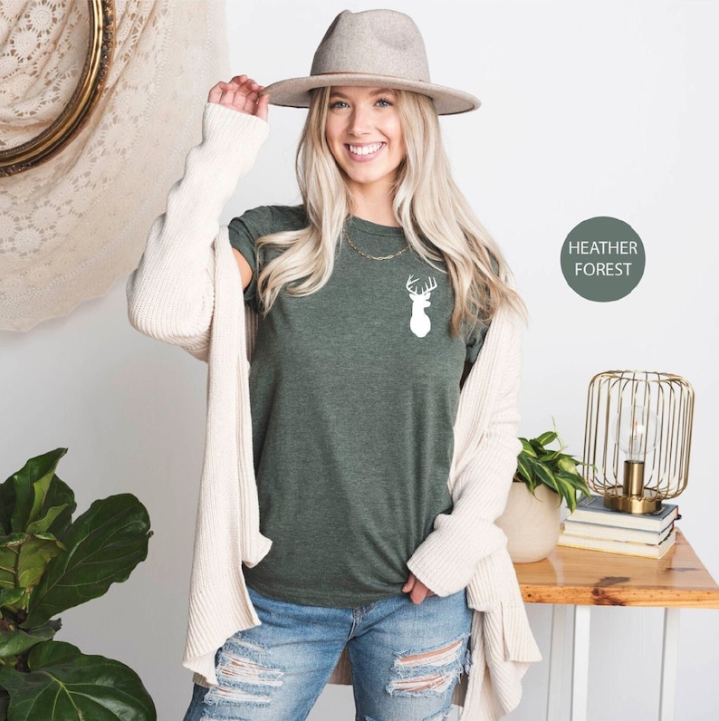 May include: A woman wearing a green t-shirt with a white deer silhouette graphic. The t-shirt is labeled "Heather Forest".