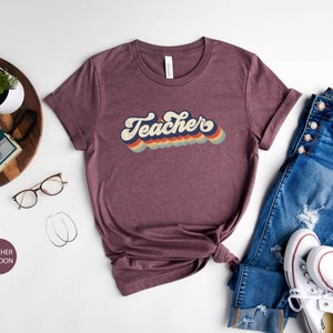 Puede incluir: Una camiseta de color marr&oacute;n rojizo con un gr&aacute;fico de estilo retro que dice "Teacher" en colores arco&iacute;ris. La camiseta est&aacute; colocada sobre una superficie blanca con un par de gafas, una bandeja de madera con una c&aacute;mara y un par de vaqueros y zapatillas blancas.