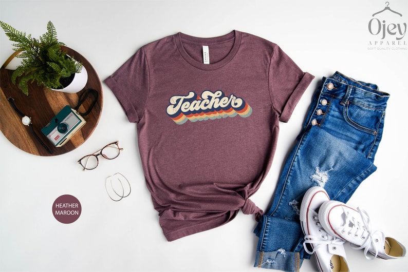 Puede incluir: Una camiseta de color marr&oacute;n rojizo con un gr&aacute;fico de estilo retro que dice "Teacher" en colores arco&iacute;ris. La camiseta est&aacute; colocada sobre una superficie blanca con un par de gafas, una bandeja de madera con una c&aacute;mara y un par de vaqueros y zapatillas blancas.