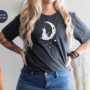 May include: A heather dark grey t-shirt with a white graphic of a cat sitting on a crescent moon with stars. The text "Ojev Apparel Soft Quality Clothing" is visible in the upper right corner of the image.
