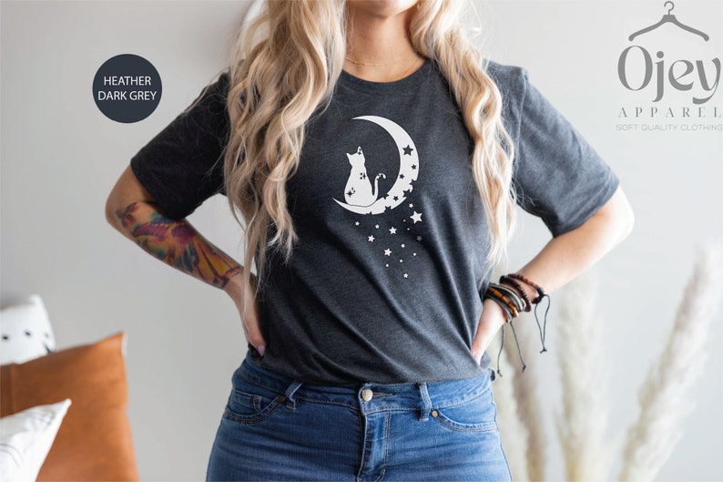 May include: A heather dark grey t-shirt with a white graphic of a cat sitting on a crescent moon with stars. The text "Ojev Apparel Soft Quality Clothing" is visible in the upper right corner of the image.
