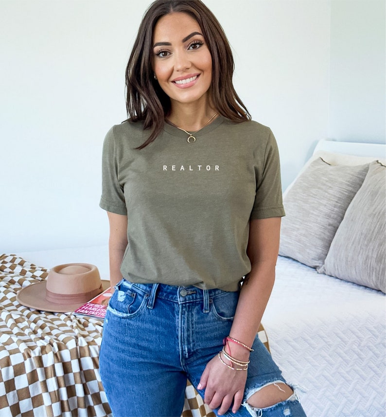 Op de afbeelding: Een vrouw draagt een groen T-shirt met het woord "REALTOR" erop gedrukt. Ze draagt ook een blauwe jeans en een ketting.