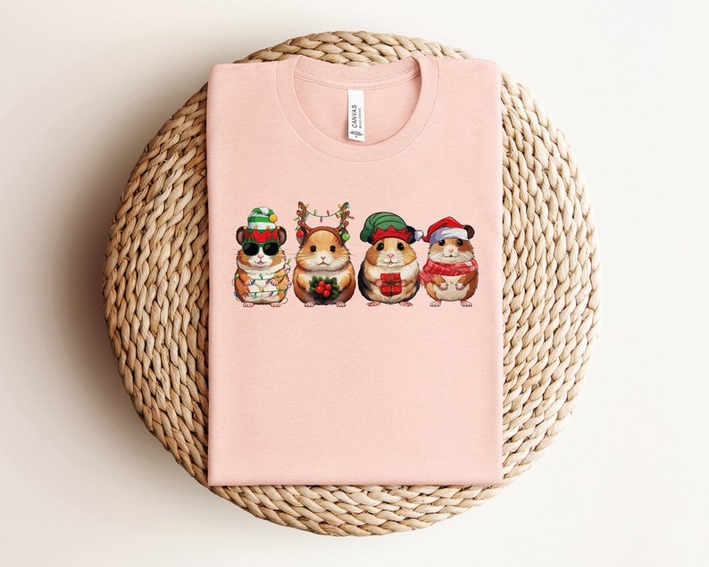 May include: Pink t-shirt with a graphic of four hamsters wearing Christmas hats and holding gifts. The hamsters are wearing Santa hats, elf hats, and reindeer antlers.