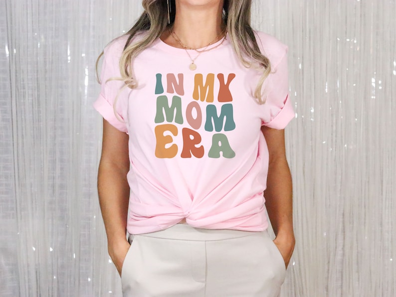 May include: A pink t-shirt with the text "In My Mom Era" in colorful, retro-style lettering.