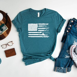 K&ouml;nnte beinhalten: Ein blaues T-Shirt mit einem wei&szlig;en, abgenutzten Design einer amerikanischen Flagge mit Angelruten und einem Fischer. Das Shirt liegt flach auf einer wei&szlig;en Oberfl&auml;che mit einer Jeans und wei&szlig;en Turnschuhen.