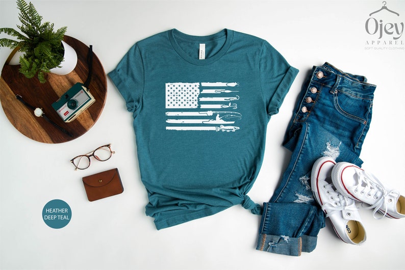 K&ouml;nnte beinhalten: Ein blaues T-Shirt mit einem wei&szlig;en, abgenutzten Design einer amerikanischen Flagge mit Angelruten und einem Fischer. Das Shirt liegt flach auf einer wei&szlig;en Oberfl&auml;che mit einer Jeans und wei&szlig;en Turnschuhen.
