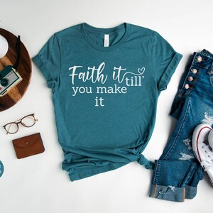 K&ouml;nnte beinhalten: Ein blaues T-Shirt mit dem Text "Faith it 'till you make it" in wei&szlig;en Buchstaben. Ein kleines Herz befindet sich neben dem Wort "till".