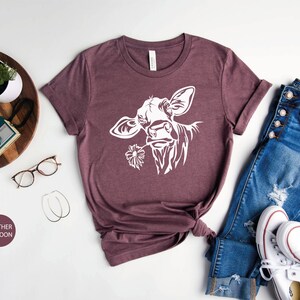 May include: A heather maroon t-shirt with a white silhouette of a cow's head with a flower in its mouth. The shirt is laid out on a white surface with a pair of jeans and white sneakers.