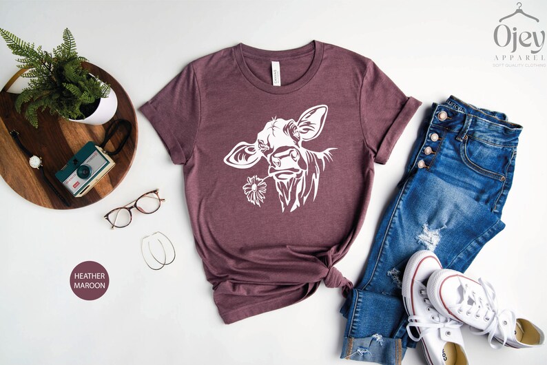 May include: A heather maroon t-shirt with a white silhouette of a cow's head with a flower in its mouth. The shirt is laid out on a white surface with a pair of jeans and white sneakers.