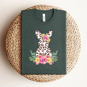 May include: A dark green t-shirt with a white and brown leopard print bunny rabbit wearing a floral crown. The bunny is surrounded by pink, yellow, and green flowers.