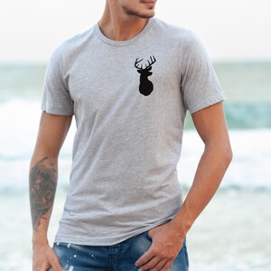 May include: A heather grey t-shirt with a black silhouette of a deer head with antlers on the front. The t-shirt is being worn by a person standing on a beach.