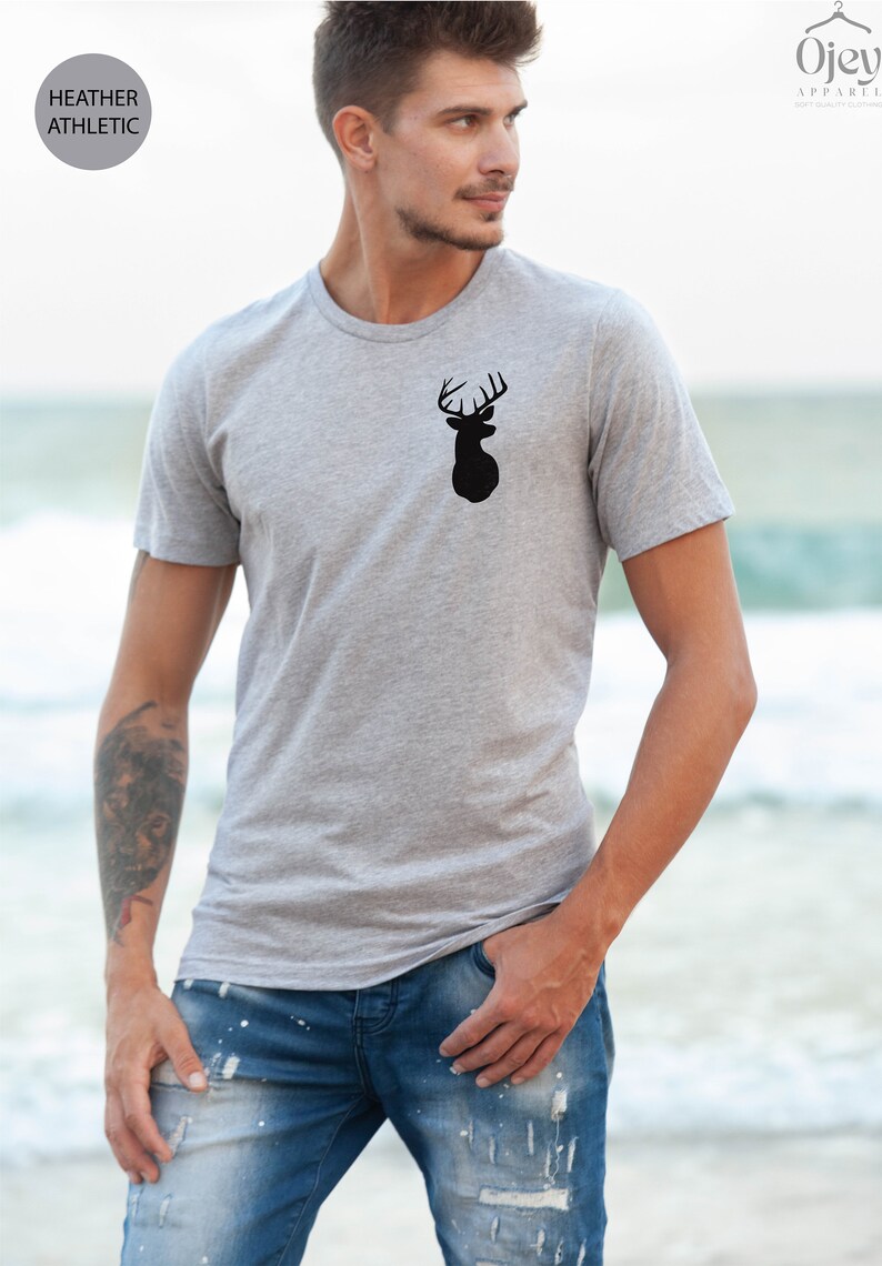 May include: A heather grey t-shirt with a black silhouette of a deer head with antlers on the front. The t-shirt is being worn by a person standing on a beach.