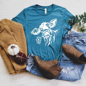 May include: A teal blue t-shirt with a white silhouette of a cow's head with a flower in its mouth. The t-shirt is laying on a white surface with a brown sweater, a pair of blue jeans, and brown boots.