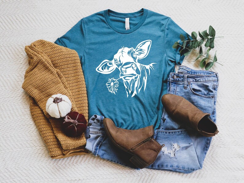 May include: A teal blue t-shirt with a white silhouette of a cow's head with a flower in its mouth. The t-shirt is laying on a white surface with a brown sweater, a pair of blue jeans, and brown boots.