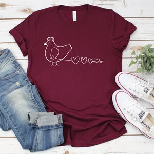 Peut inclure: Un t-shirt de couleur bordeaux avec un motif blanc repr&eacute;sentant un poulet suivi d'une cha&icirc;ne de c&oelig;urs.
