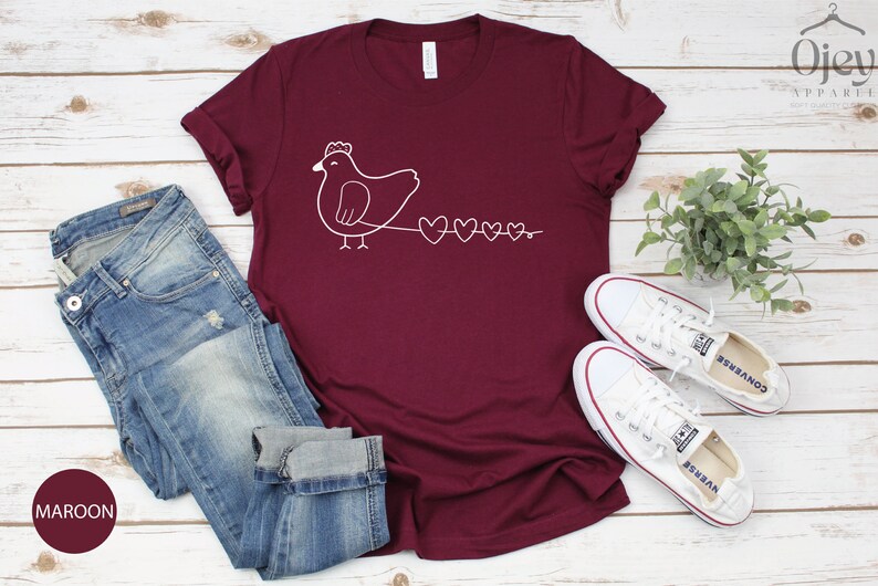 Peut inclure: Un t-shirt de couleur bordeaux avec un motif blanc repr&eacute;sentant un poulet suivi d'une cha&icirc;ne de c&oelig;urs.