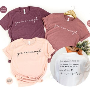 May include: Three t-shirts with the text "you are enough" in white script on a maroon, peach, and mauve background. The back of a white t-shirt has the text "Dear person behind me: The world is a better place with you in it. Lots of love, - the person in front of you".