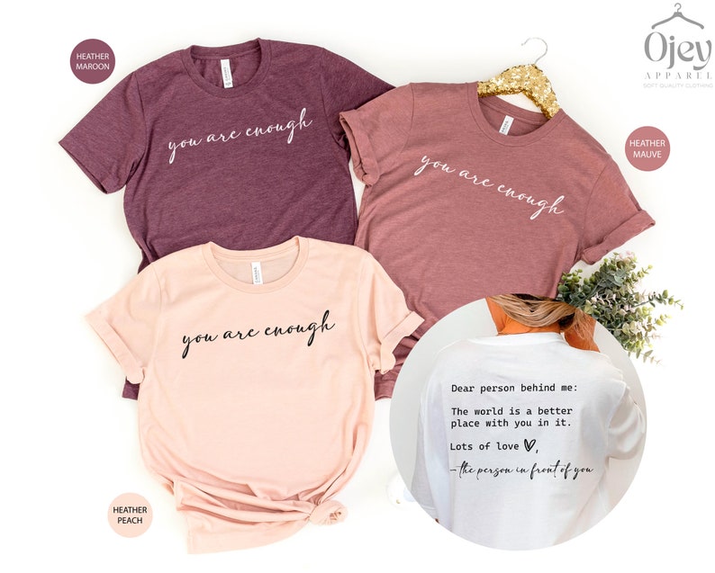 May include: Three t-shirts with the text "you are enough" in white script on a maroon, peach, and mauve background. The back of a white t-shirt has the text "Dear person behind me: The world is a better place with you in it. Lots of love, - the person in front of you".