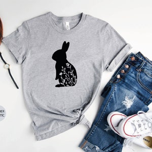 May include: A heather gray t-shirt with a black silhouette of a rabbit with floral details. The shirt is shown on a white background with a pair of jeans and white sneakers.