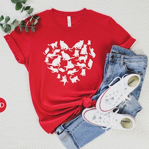 May include: Red t-shirt with a white heart design made up of multiple cat silhouettes. The shirt is laid flat on a white surface with a pair of white sneakers and blue jeans.