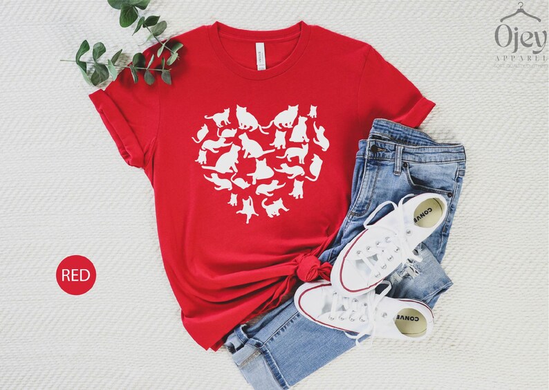 May include: Red t-shirt with a white heart design made up of multiple cat silhouettes. The shirt is laid flat on a white surface with a pair of white sneakers and blue jeans.