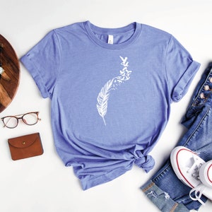 Op de afbeelding: Een heather columbia blauw t-shirt met een witte afbeelding van een veer met vogels die eruit vliegen. Het shirt ligt op een wit oppervlak met een bril, een bruine portemonnee, een houten dienblad met een camera en een horloge, en een paar witte sneakers en een blauwe jeans.