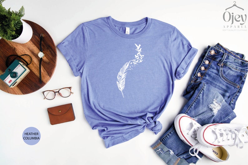 Op de afbeelding: Een heather columbia blauw t-shirt met een witte afbeelding van een veer met vogels die eruit vliegen. Het shirt ligt op een wit oppervlak met een bril, een bruine portemonnee, een houten dienblad met een camera en een horloge, en een paar witte sneakers en een blauwe jeans.
