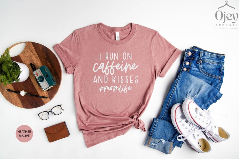 K&ouml;nnte beinhalten: Ein T-Shirt in Heide-Mauve mit dem Aufdruck "I run on caffeine and kisses #momlife" in Wei&szlig;.