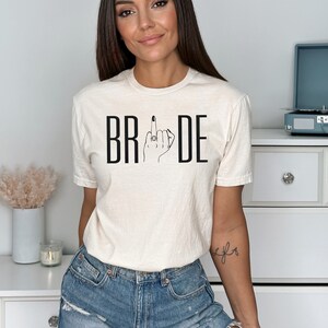 May include: A white t-shirt with the word "BRIDE" printed in black letters. The "I" in bride is replaced with a black and white illustration of a hand giving the middle finger with a ring on the ring finger.