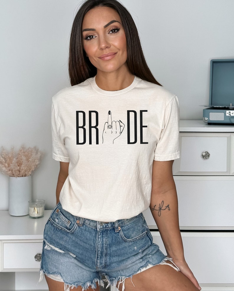 May include: A white t-shirt with the word "BRIDE" printed in black letters. The "I" in bride is replaced with a black and white illustration of a hand giving the middle finger with a ring on the ring finger.