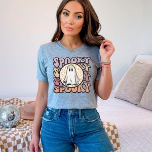 Puede incluir: Una camiseta gris con un gr&aacute;fico de fantasma y el texto "SPOOKY" en una fuente retro. El fantasma est&aacute; en un c&iacute;rculo con un fondo de estampado de leopardo.