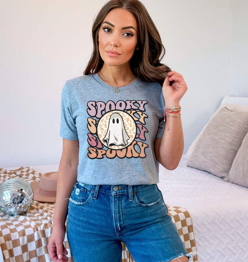 Puede incluir: Una camiseta gris con un gr&aacute;fico de fantasma y el texto "SPOOKY" en una fuente retro. El fantasma est&aacute; en un c&iacute;rculo con un fondo de estampado de leopardo.