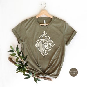 May include: A green t-shirt with a white graphic of a desert scene with cacti, a sun, and mountains. The text "HEATHER OLIVE" is visible in the bottom right corner of the image.