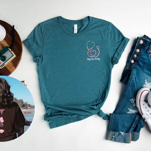 May include: Teal blue t-shirt with a pink stethoscope and heart graphic and the text "Nurse-tastic". The shirt is laid flat on a white surface with a pair of blue jeans and white sneakers.