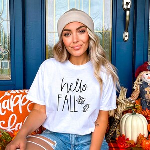 May include: A white t-shirt with the text "hello fall" in black script lettering and a small leaf design. The model is wearing the shirt with a beige beanie and denim shorts.