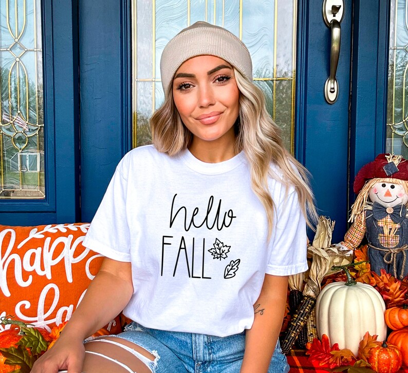 May include: A white t-shirt with the text "hello fall" in black script lettering and a small leaf design. The model is wearing the shirt with a beige beanie and denim shorts.