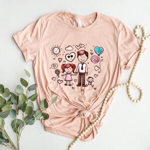 Peut inclure: Un t-shirt rose p&acirc;le avec une illustration dessin&eacute;e &agrave; la main d'une famille. L'illustration repr&eacute;sente un p&egrave;re, une fille et un b&eacute;b&eacute;. Le p&egrave;re tient la main de sa fille. L'illustration est entour&eacute;e de c&oelig;urs, d'un soleil, d'un nuage et de sucettes.