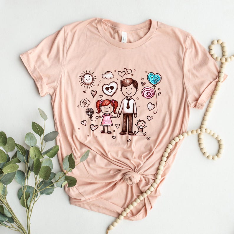 Peut inclure: Un t-shirt rose p&acirc;le avec une illustration dessin&eacute;e &agrave; la main d'une famille. L'illustration repr&eacute;sente un p&egrave;re, une fille et un b&eacute;b&eacute;. Le p&egrave;re tient la main de sa fille. L'illustration est entour&eacute;e de c&oelig;urs, d'un soleil, d'un nuage et de sucettes.
