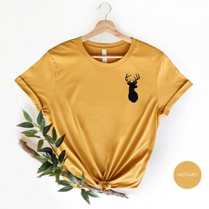 May include: A mustard yellow t-shirt with a black silhouette of a deer head on the left side of the chest. The shirt is on a hanger and has a knotted bottom.